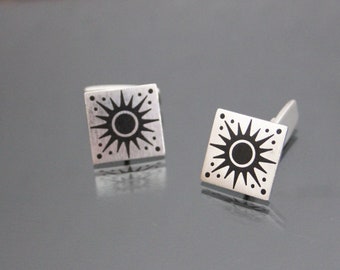 Puede incluir: Un par de gemelos de plata con un diseño de sol negro. El sol tiene un círculo en el centro y rayos que se extienden hacia afuera.