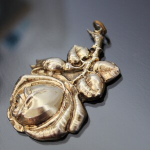 May include: A silver-toned metal pendant in the shape of a rose with leaves. The pendant has a detailed, three-dimensional design, with the rose petals and leaves clearly defined. The background is a dark, reflective surface.