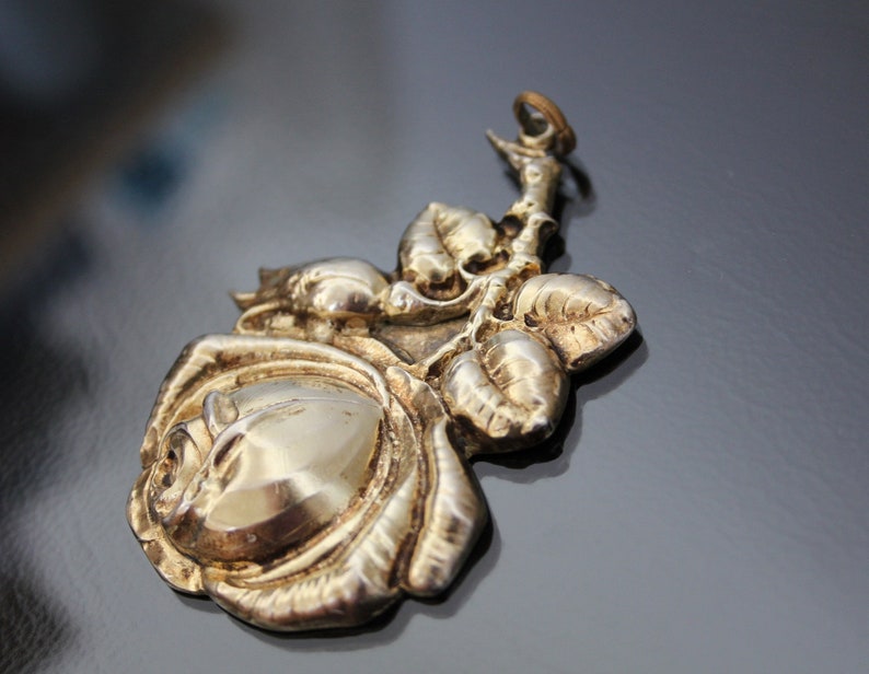 May include: A silver-toned metal pendant in the shape of a rose with leaves. The pendant has a detailed, three-dimensional design, with the rose petals and leaves clearly defined. The background is a dark, reflective surface.