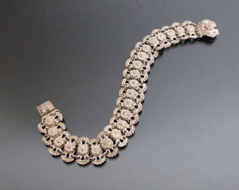 Puede incluir: Un brazalete de plata con un diseño delicado e intrincado. El brazalete está formado por una serie de pequeños eslabones redondos con forma de flor.