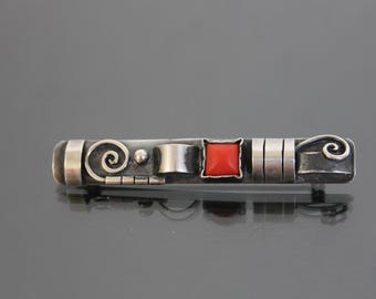 May include: A silver brooch with a red stone and intricate silver designs. The brooch has a swirl design on one end and a leaf design on the other end.