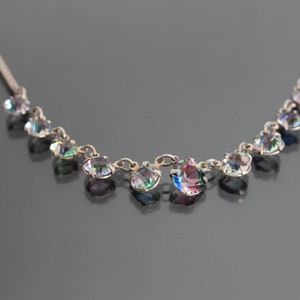 Pode incluir: Um colar de prata com pedras preciosas redondas e iridescentes. As pedras estão dispostas em uma linha curva, conectadas por uma corrente fina. As pedras refletem um arco-íris de cores, incluindo rosa, verde e azul.