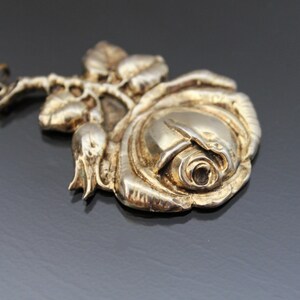 May include: A silver-toned rose pendant with detailed petals, leaves, and a stem. The pendant has a vintage look and is likely made of metal. The rose is the focal point, with intricate detailing on the petals and leaves.