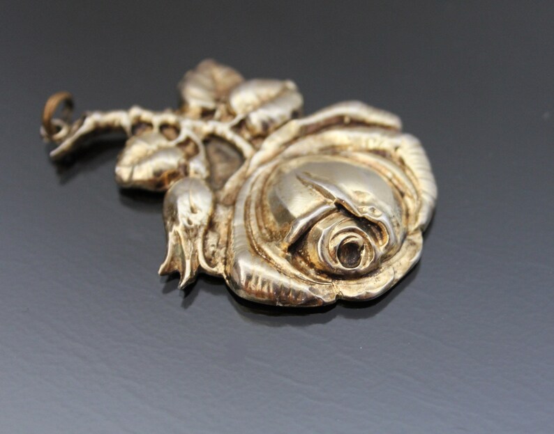 May include: A silver-toned rose pendant with detailed petals, leaves, and a stem. The pendant has a vintage look and is likely made of metal. The rose is the focal point, with intricate detailing on the petals and leaves.