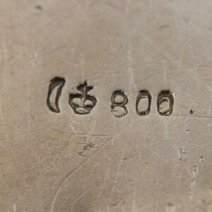 May include: Close-up of a silver metal surface with stamped markings. The markings include a crescent moon shape, a crown, and the number "800". The surface has visible scratches and imperfections.