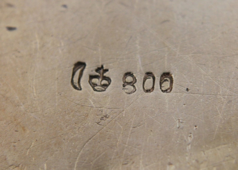 May include: Close-up of a silver metal surface with stamped markings. The markings include a crescent moon shape, a crown, and the number "800". The surface has visible scratches and imperfections.
