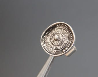 May include: A silver ring with a circular design featuring concentric circles and a small raised dot in the centre.
