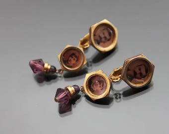 May include: A pair of gold-toned earrings with a purple crystal dangle. Each earring features a round, gold-toned locket with a black and white photo of a person inside.