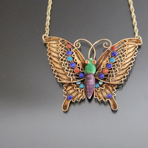 Può includere: Una collana a forma di farfalla in oro con accenti di smalto colorati. La farfalla è realizzata in filo di filigrana e presenta un design dettagliato. La collana è un gioiello bellissimo e unico.