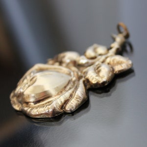 May include: A detailed, antique-style pendant in a gold tone, featuring a rose design. The pendant has a central oval shape and is surrounded by sculpted leaves and petals. The bail is a small loop for attaching to a chain.