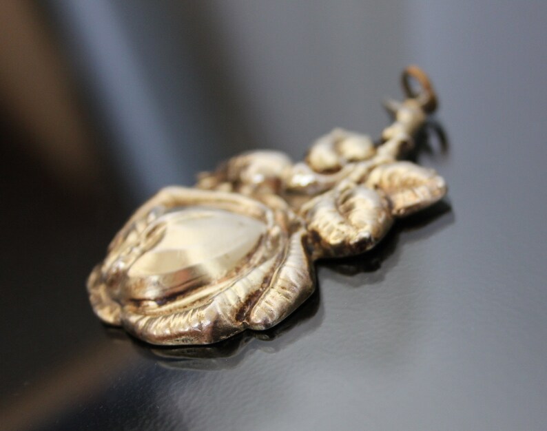 May include: A detailed, antique-style pendant in a gold tone, featuring a rose design. The pendant has a central oval shape and is surrounded by sculpted leaves and petals. The bail is a small loop for attaching to a chain.