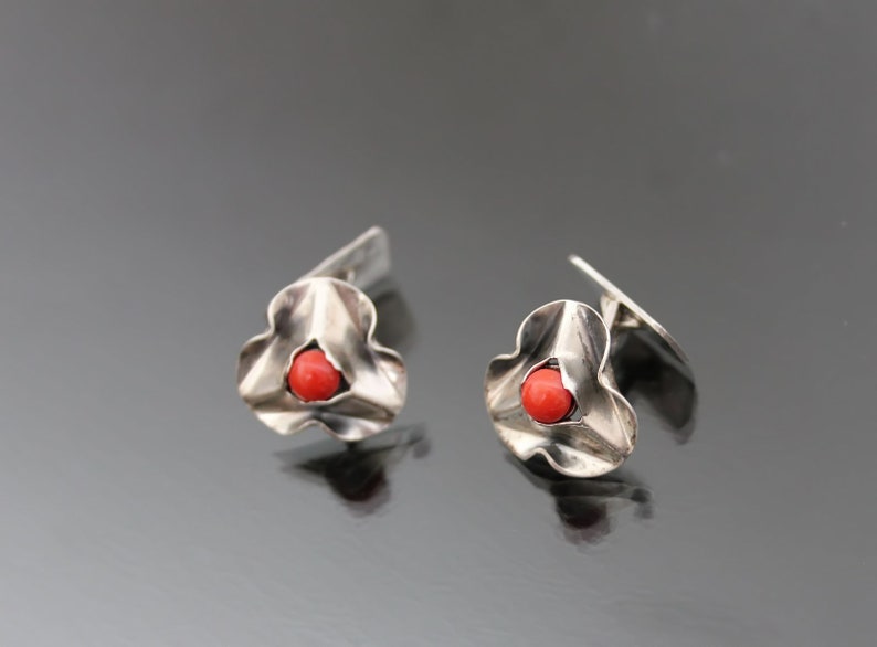 Puede incluir: Un par de gemelos de plata con una piedra de coral rojo en el centro de cada gemelo. Los gemelos tienen forma de flor con cinco p&eacute;talos.