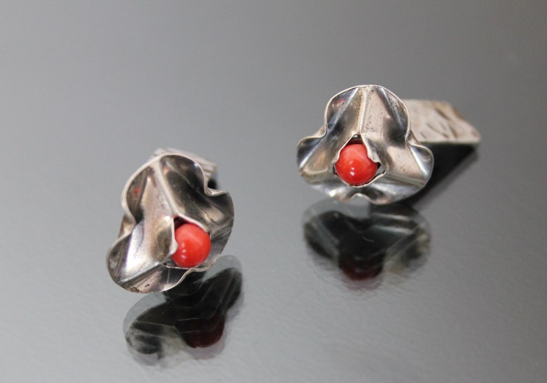 Puede incluir: Un par de pendientes de plata con una piedra de coral rojo en el centro de cada pendiente. Los pendientes tienen forma de flor con una superficie texturizada.