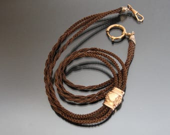 Puede incluir: Una cadena de reloj trenzada marrón con detalles dorados y un dije en forma de corazón.