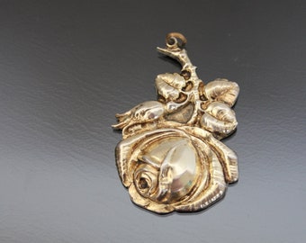 Può includere: Un ciondolo color argento a forma di rosa. Il ciondolo presenta un bocciolo di rosa dettagliato con diverse foglie e uno stelo. Il metallo ha una finitura lucida, che riflette la luce. Il ciondolo è probabilmente per una collana o altri gioielli.