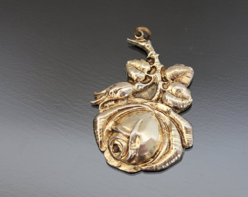 May include: A silver-toned pendant in the shape of a rose. The pendant features a detailed rose bloom with several leaves and a stem. The metal has a polished finish, reflecting light. The pendant is likely for a necklace or other jewelry.