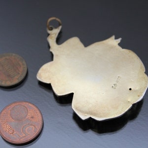 May include: A silver-colored pendant with a floral design, featuring a loop at the top for hanging. Two coins, a penny and a nickel, are next to the pendant. The pendant has a small hole and the text "800".