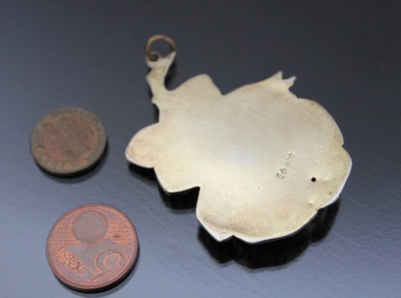 May include: A silver-colored pendant with a floral design, featuring a loop at the top for hanging. Two coins, a penny and a nickel, are next to the pendant. The pendant has a small hole and the text "800".