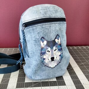May include: A small denim backpack with a geometric wolf design on the front. The backpack has a zipper closure and a dark blue adjustable strap.
