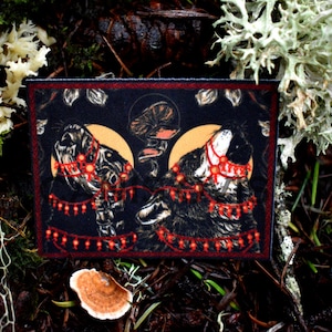 May include: A black and white illustration of two dogs wearing red and gold necklaces with dangling ornaments. The dogs are set against a dark background with a crescent moon behind each dog.