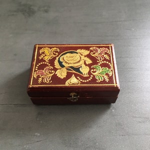 May include: A small, red wooden box with a gold-painted floral design. The box has a hinged lid and a small metal clasp.