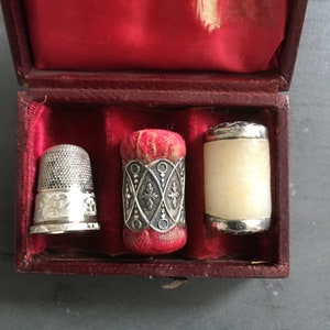 May include: A red velvet box with three vintage thimbles. One thimble is silver with an intricate design, another is silver with a white top, and the third is red with silver trim.