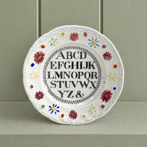 May include: A white ceramic plate featuring a central black alphabet from A to Z followed by an ampersand, enclosed within a decorative border. The rim is adorned with a colourful pattern of small painted flowers in red, yellow, blue, and green.