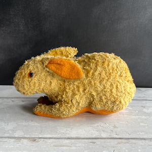 May include: A vintage plush rabbit toy in a seated position. The rabbit has a textured, golden-yellow fur body with orange inner ears and feet. It has small, red eyes and is set against a dark background.