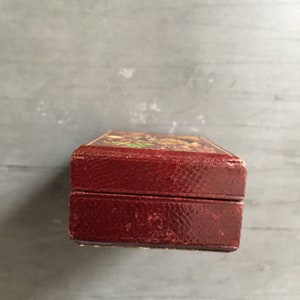 May include: A small, rectangular, red leather box with a gold-colored design on the top. The box is closed and the lid is slightly ajar.