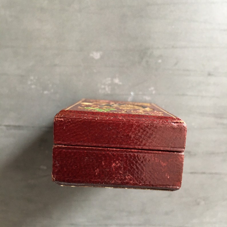 May include: A small, rectangular, red leather box with a gold-colored design on the top. The box is closed and the lid is slightly ajar.