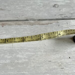 May include: A vintage measuring tape with a cream-coloured tape and black markings. The tape extends from a small, cylindrical case with blue and white stripes. The case has a metal ring and a brass-coloured rim. The tape measures up to 10 cm.