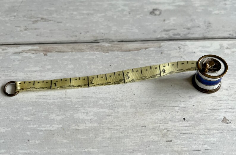 May include: A vintage measuring tape with a cream-coloured tape and black markings. The tape extends from a small, cylindrical case with blue and white stripes. The case has a metal ring and a brass-coloured rim. The tape measures up to 10 cm.