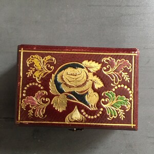 May include: A small, red, rectangular box with a gold and black floral design. The box has a hinged lid and a small metal clasp.