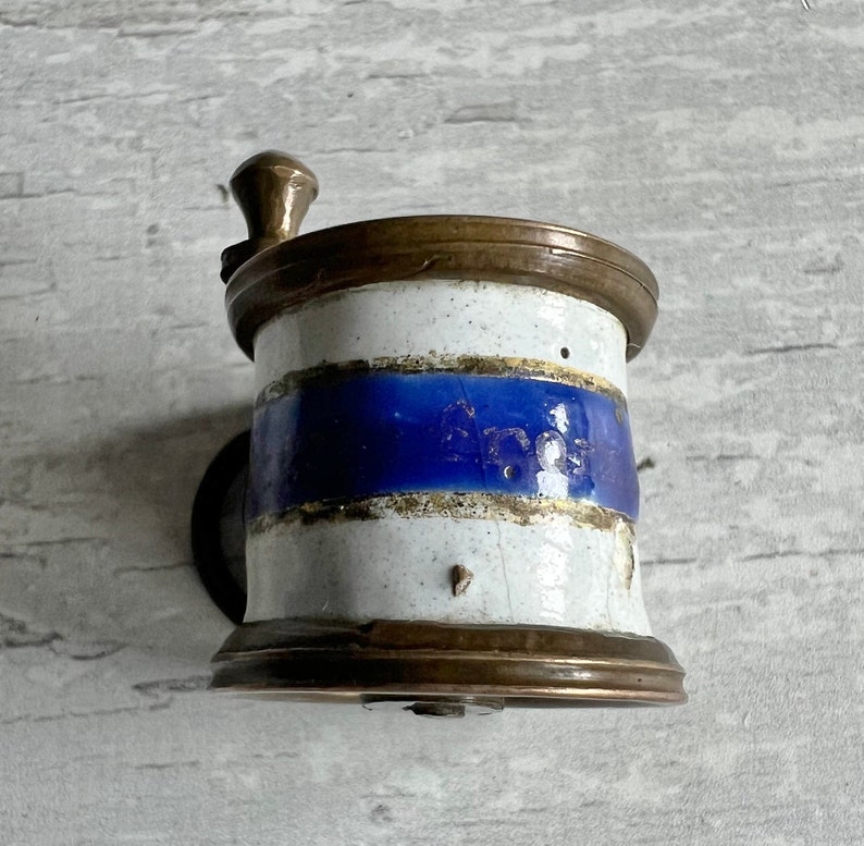 May include: Vintage coffee grinder with a white and blue ceramic body, complemented by brass fittings. The grinder has a hand crank and a cylindrical form, displaying a classic appearance. The blue band provides a striking contrast to the design.