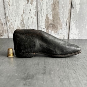 May include: A black leather shoe form, also known as a shoe last, used for shaping shoes. The form is made of leather and has a rounded toe. A small gold thimble is in the foreground.
