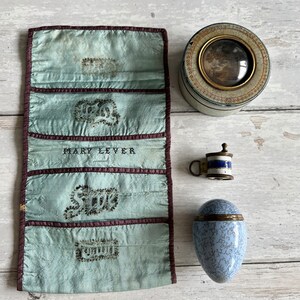 May include: A collection of vintage objects: a pale blue silk needle case with embroidered details, a small circular tin with a framed picture, a miniature mug, and a blue and white egg-shaped container.