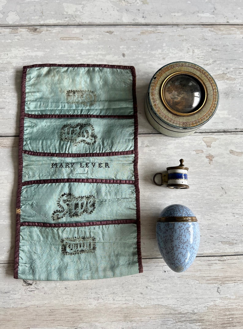 May include: A collection of vintage objects: a pale blue silk needle case with embroidered details, a small circular tin with a framed picture, a miniature mug, and a blue and white egg-shaped container.