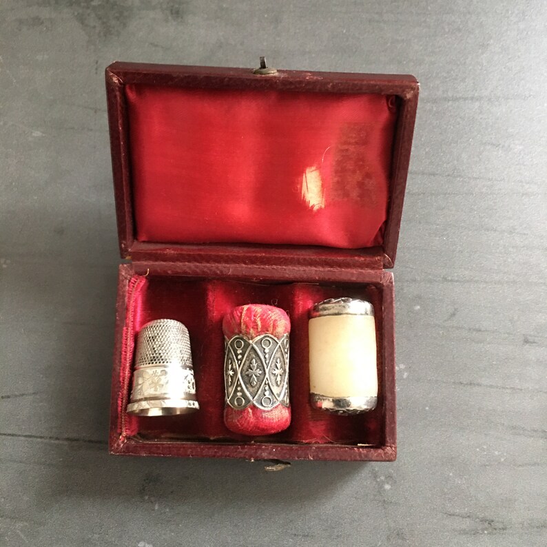 May include: A red velvet box with three silver thimbles. The thimbles are decorated with intricate designs. One thimble is red and white, another is white, and the third is silver.