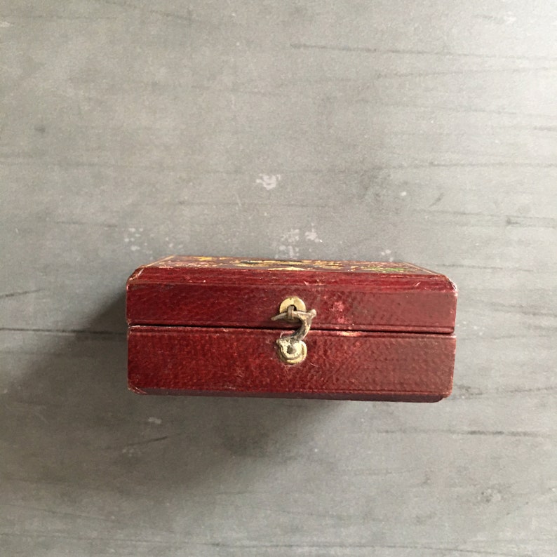 May include: A small, red, wooden box with a gold latch. The box has a textured surface and is closed.