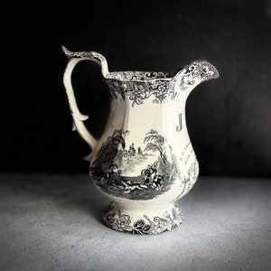 May include: A white ceramic pitcher with a black and white floral design. The pitcher has a handle and a spout. The design includes a hunting scene with a dog, a horse, and a rider.