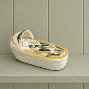 May include: A white ceramic cradle figurine featuring a yellow trim along the hood and edges. The interior of the cradle is decorated with a painted pattern of green and dark brown abstract shapes on a white background, creating a textured appearance.