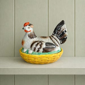 Peut inclure: Figurine de poule en céramique avec une crête rouge, un corps tacheté blanc et marron, et des plumes de queue noires et blanches. La poule est posée sur un socle en forme de panier jaune. Objet de décoration de cuisine.