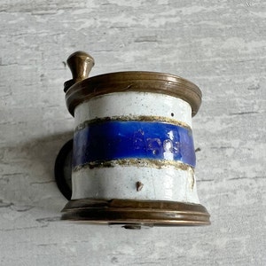 May include: Vintage coffee grinder with a white and blue ceramic body and brass details. The grinder has a hand crank and a small, round base. The ceramic has a blue stripe and brass rims.