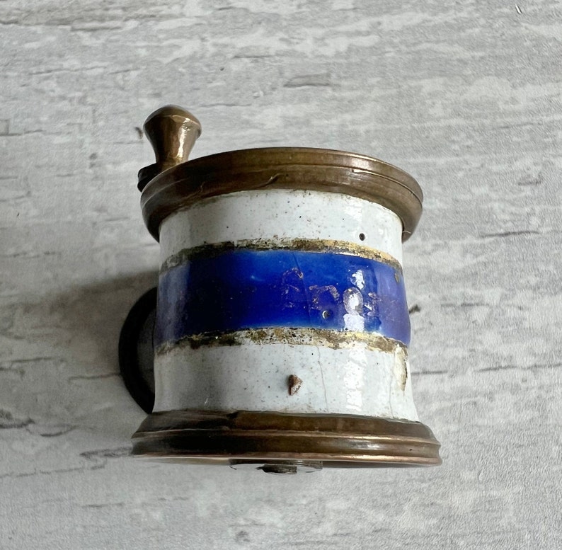 May include: Vintage coffee grinder with a white and blue ceramic body and brass details. The grinder has a hand crank and a small, round base. The ceramic has a blue stripe and brass rims.
