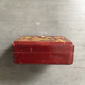 May include: A small, red leather box with gold accents and a decorative design on the top. The box is closed and the lid is not visible.