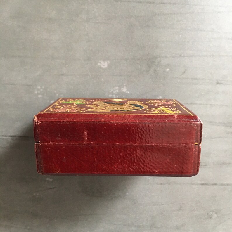 May include: A small, red leather box with gold accents and a decorative design on the top. The box is closed and the lid is not visible.