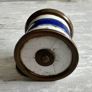 May include: A cylindrical bottle stopper with a white base, a blue stripe, and bronze-coloured metal rims. The stopper has a circular base with a central metal disc and a small screw. The design is reminiscent of vintage decor.