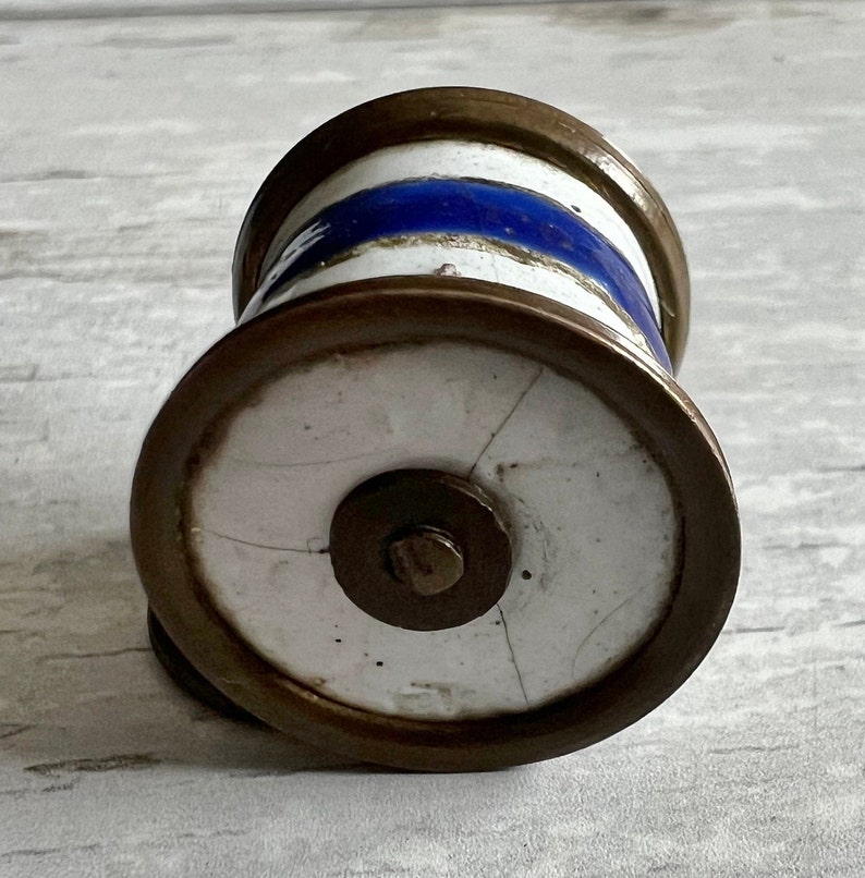 May include: A cylindrical bottle stopper with a white base, a blue stripe, and bronze-coloured metal rims. The stopper has a circular base with a central metal disc and a small screw. The design is reminiscent of vintage decor.