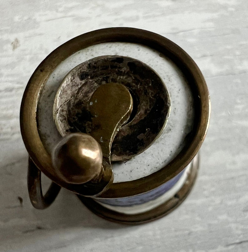 May include: Close-up of a vintage sewing thread spool. The spool features a brass-coloured metal frame with a white and blue ceramic design. A brass-coloured metal lever and a small ball are attached to the spool. The thread is a light blue colour.