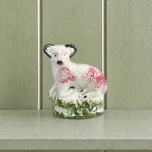 May include: A ceramic figurine of a sheep with a white body, black ears, and red speckled markings on its back and sides. The sheep stands on a textured green base that resembles grass, featuring a glossy finish and hand-painted details.